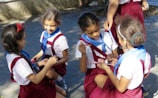 Several young children are sitting and kneeling on the ground, wearing uniforms with white shirts, red skirts, and blue scarves. They appear to be engaged in an activity, possibly drawing or writing, as they hold markers or pens in their hands. They are outside, and the ground is paved, with shadows indicating sunlight.