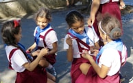 Several young children are sitting and kneeling on the ground, wearing uniforms with white shirts, red skirts, and blue scarves. They appear to be engaged in an activity, possibly drawing or writing, as they hold markers or pens in their hands. They are outside, and the ground is paved, with shadows indicating sunlight.