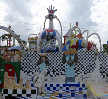 A colorful and eclectic mosaic installation features abstract and surreal elements. It includes a large structure with a hand and eye motif at the top, checkerboard patterns, and stylized human figures with intense facial expressions. The artwork is vibrant with a mix of geometric shapes, flowing forms, and varied textures.