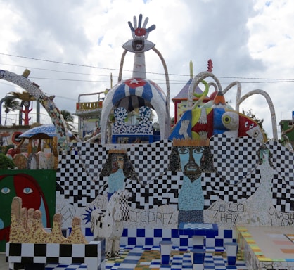 A colorful and eclectic mosaic installation features abstract and surreal elements. It includes a large structure with a hand and eye motif at the top, checkerboard patterns, and stylized human figures with intense facial expressions. The artwork is vibrant with a mix of geometric shapes, flowing forms, and varied textures.