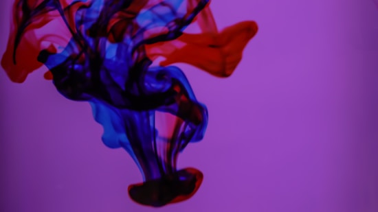 Vibrant ink flows and swirls underwater, blending striking shades of blue and red against a purple background. The colors create a mesmerizing, fluid pattern reminiscent of abstract art.