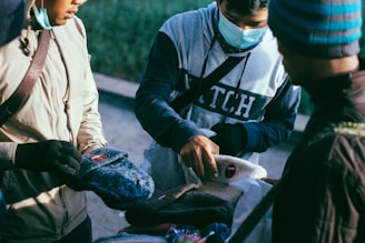 Several individuals are gathered around looking at various knitted hats. One person is pointing at a beanie while wearing a face mask and gloves. The setting appears to be outdoors with some greenery in the background.