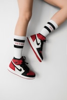 A pair of legs wearing high-top sneakers in red, black, and white colors paired with white socks that have 'Vetements' written in red. The socks also have black stripes near the top.