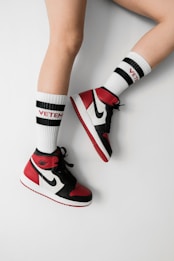 A pair of legs wearing high-top sneakers in red, black, and white colors paired with white socks that have 'Vetements' written in red. The socks also have black stripes near the top.