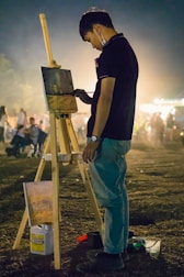 A person is intently painting on a canvas set on a wooden easel outdoors at night. They are standing on a grassy area surrounded by other people in the background, which are out of focus. The painter is wearing a dark shirt and light pants, with a mask hanging from one ear. The surroundings are dimly lit, creating an atmospheric scene.