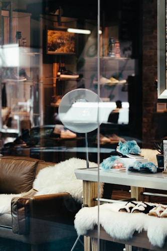 A stylish shop window displays plush furniture and accessories. A comfortable brown leather couch is adorned with fluffy white throws, creating a cozy atmosphere. On a wooden table nearby are soft, blue-hued slippers and a furry blanket. The background includes shelves showcasing a variety of items, illuminated softly by overhead lights.