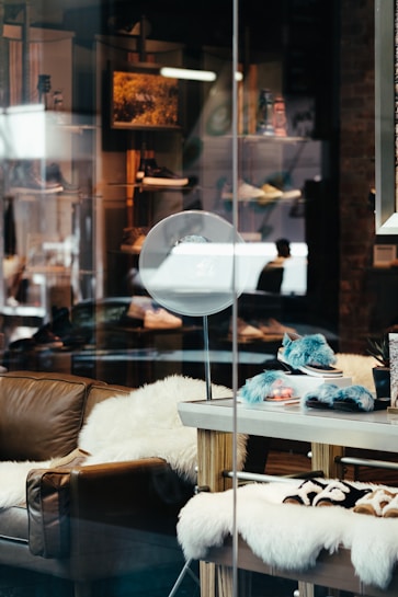 A stylish shop window displays plush furniture and accessories. A comfortable brown leather couch is adorned with fluffy white throws, creating a cozy atmosphere. On a wooden table nearby are soft, blue-hued slippers and a furry blanket. The background includes shelves showcasing a variety of items, illuminated softly by overhead lights.