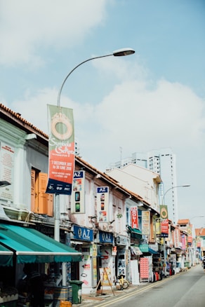 A lively street scene with colorful shopfronts featuring various signs and advertisements. The buildings have a traditional architectural style with white walls and orange-tiled roofs. A tall, modern building can be seen in the background, while a street lamp and banners hang above the street.