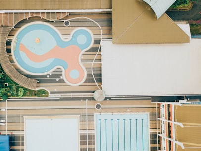 Aerial view of two swimming pools, one with an irregular, playful shape featuring a mix of blue and orange colors, and another rectangular pool with lanes marked for swimming. The pools are surrounded by deck areas and landscaping, featuring greenery and structured pathways.