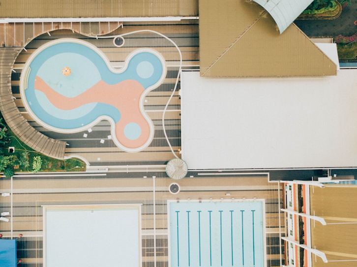 Aerial view of two swimming pools, one with an irregular, playful shape featuring a mix of blue and orange colors, and another rectangular pool with lanes marked for swimming. The pools are surrounded by deck areas and landscaping, featuring greenery and structured pathways.