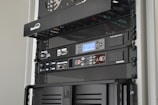 A server rack containing various network and power management devices. The devices feature buttons, switches, and digital displays, indicating active usage and monitoring capabilities. The rack has a black metal mesh design for ventilation.