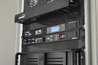 A server rack containing various network and power management devices. The devices feature buttons, switches, and digital displays, indicating active usage and monitoring capabilities. The rack has a black metal mesh design for ventilation.