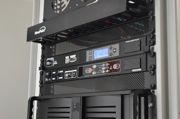 A server rack containing various network and power management devices. The devices feature buttons, switches, and digital displays, indicating active usage and monitoring capabilities. The rack has a black metal mesh design for ventilation.