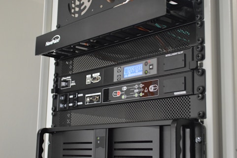 A server rack containing various network and power management devices. The devices feature buttons, switches, and digital displays, indicating active usage and monitoring capabilities. The rack has a black metal mesh design for ventilation.