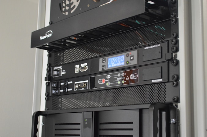 A server rack containing various network and power management devices. The devices feature buttons, switches, and digital displays, indicating active usage and monitoring capabilities. The rack has a black metal mesh design for ventilation.