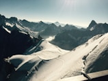 A vast and majestic mountain landscape with snow-covered peaks and rugged ridges. A group of people are seen trekking along a narrow snow trail, dwarfed by the grand scale of the surrounding scenery. The horizon features a range of high mountains under a clear blue sky.