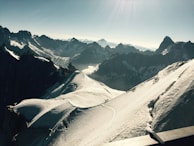 A vast and majestic mountain landscape with snow-covered peaks and rugged ridges. A group of people are seen trekking along a narrow snow trail, dwarfed by the grand scale of the surrounding scenery. The horizon features a range of high mountains under a clear blue sky.