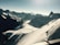 A vast and majestic mountain landscape with snow-covered peaks and rugged ridges. A group of people are seen trekking along a narrow snow trail, dwarfed by the grand scale of the surrounding scenery. The horizon features a range of high mountains under a clear blue sky.