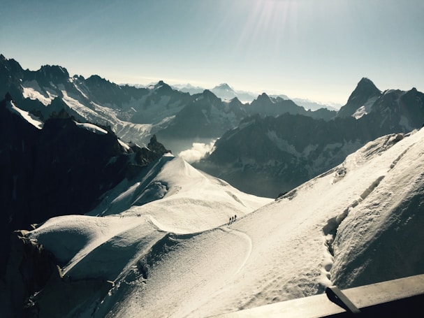 A vast and majestic mountain landscape with snow-covered peaks and rugged ridges. A group of people are seen trekking along a narrow snow trail, dwarfed by the grand scale of the surrounding scenery. The horizon features a range of high mountains under a clear blue sky.