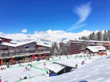 A snowy alpine resort with modern buildings and wooden lodges, surrounded by snow-covered mountains and evergreen trees. People are engaging in winter activities on the ski slopes in bright sunlight under a clear blue sky.