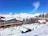 A snowy alpine resort with modern buildings and wooden lodges, surrounded by snow-covered mountains and evergreen trees. People are engaging in winter activities on the ski slopes in bright sunlight under a clear blue sky.