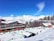 A snowy alpine resort with modern buildings and wooden lodges, surrounded by snow-covered mountains and evergreen trees. People are engaging in winter activities on the ski slopes in bright sunlight under a clear blue sky.