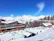 A snowy alpine resort with modern buildings and wooden lodges, surrounded by snow-covered mountains and evergreen trees. People are engaging in winter activities on the ski slopes in bright sunlight under a clear blue sky.