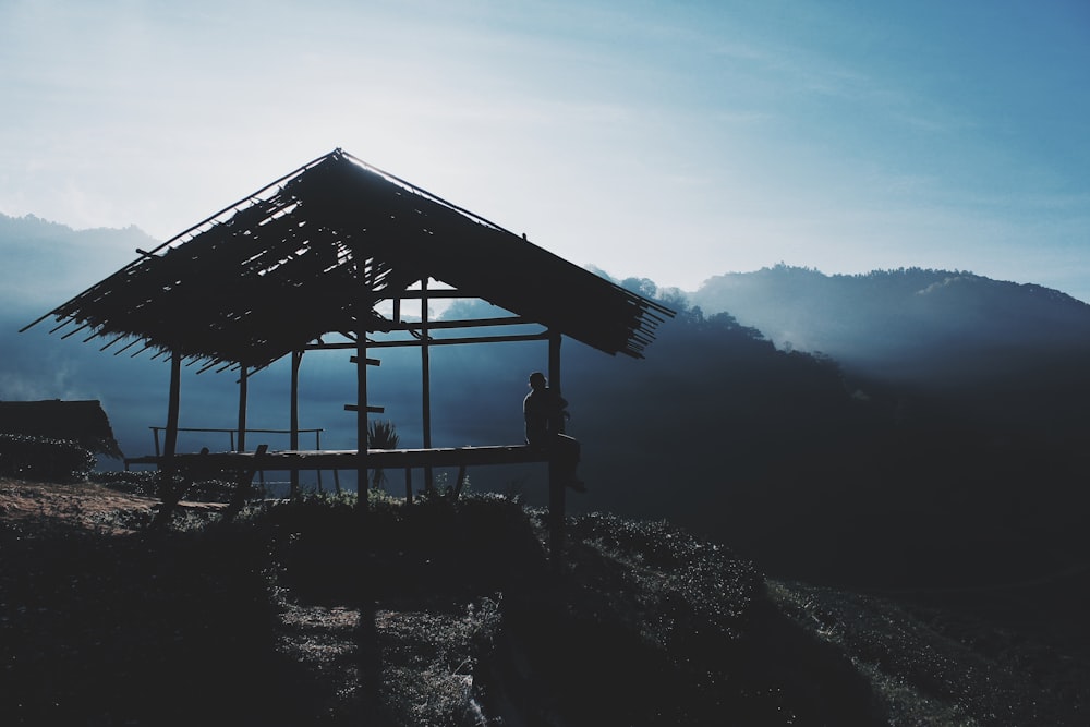 silhouette photo of person sitting on nippa hut on hill in front of mountains surrounded by fogs