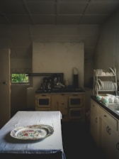 A vintage kitchen setting with an old-fashioned stove and cabinets, a table covered with a white cloth, and a decorative platter on top. Plates and cups are neatly arranged on a dish rack on the right side. The lighting is dim, adding to an antique ambiance.