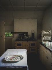 A vintage kitchen setting with an old-fashioned stove and cabinets, a table covered with a white cloth, and a decorative platter on top. Plates and cups are neatly arranged on a dish rack on the right side. The lighting is dim, adding to an antique ambiance.
