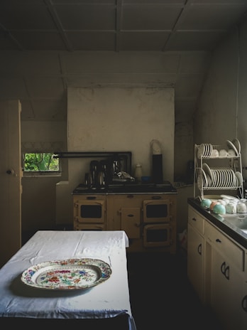 A vintage kitchen setting with an old-fashioned stove and cabinets, a table covered with a white cloth, and a decorative platter on top. Plates and cups are neatly arranged on a dish rack on the right side. The lighting is dim, adding to an antique ambiance.