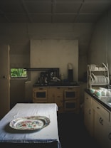 A vintage kitchen setting with an old-fashioned stove and cabinets, a table covered with a white cloth, and a decorative platter on top. Plates and cups are neatly arranged on a dish rack on the right side. The lighting is dim, adding to an antique ambiance.