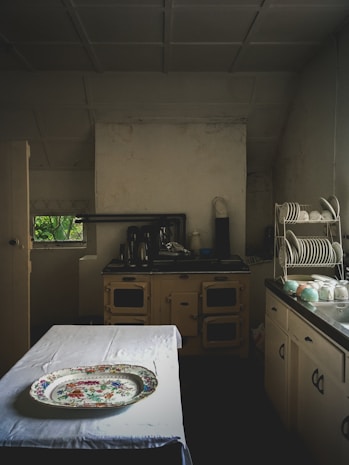 A vintage kitchen setting with an old-fashioned stove and cabinets, a table covered with a white cloth, and a decorative platter on top. Plates and cups are neatly arranged on a dish rack on the right side. The lighting is dim, adding to an antique ambiance.