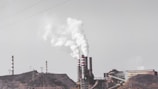 A factory complex with large chimneys emitting thick white smoke into a gray sky. The industrial setting includes multiple buildings and power lines, set against a barren landscape.