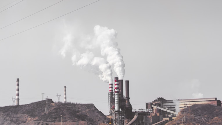 A factory complex with large chimneys emitting thick white smoke into a gray sky. The industrial setting includes multiple buildings and power lines, set against a barren landscape.
