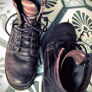 A pair of well-worn brown leather boots with black laces is placed on a decorative tile floor featuring a floral pattern. The boots show signs of use and have a rustic, vintage appearance.