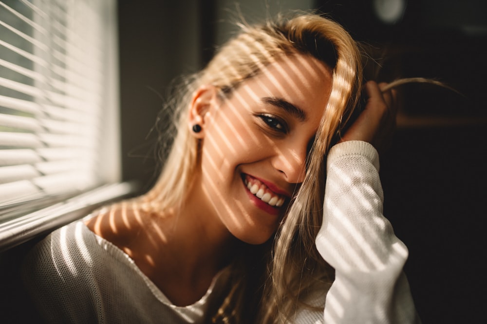 selective focus photography of smiling woman holding her hair beside window blinds