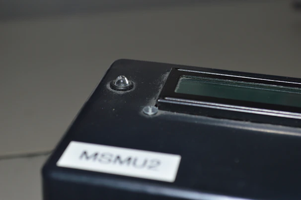A close-up of a black electronic device with a small screen and a label marked 'MSMU2'. The surface appears slightly dusty, and there are two small protruding knobs or fasteners.