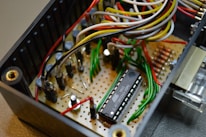 An electronic circuit featuring a microcontroller mounted on a perforated board with various wires in multiple colors connecting to different components. The setup includes resistors, capacitors, and other small electronic parts.