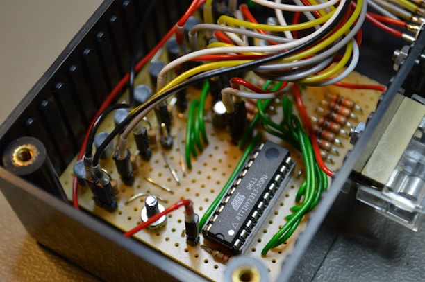 An electronic circuit featuring a microcontroller mounted on a perforated board with various wires in multiple colors connecting to different components. The setup includes resistors, capacitors, and other small electronic parts.