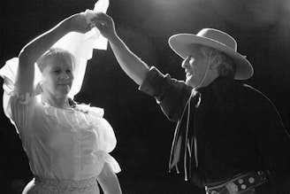A man and woman are dancing together, with the woman holding a flowing white cloth above them. The man is wearing a broad-brimmed hat and traditional attire, while the woman has on a ruffled blouse. They appear joyful, captured in a moment of movement on stage under dramatic lighting.