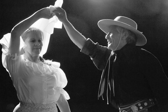 A man and woman are dancing together, with the woman holding a flowing white cloth above them. The man is wearing a broad-brimmed hat and traditional attire, while the woman has on a ruffled blouse. They appear joyful, captured in a moment of movement on stage under dramatic lighting.