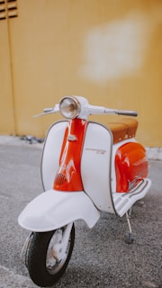 A vintage-style scooter with a vibrant orange and white color scheme is parked on a gray asphalt surface. It features a classic round headlight and chrome handlebars, set against a mustard-yellow wall.
