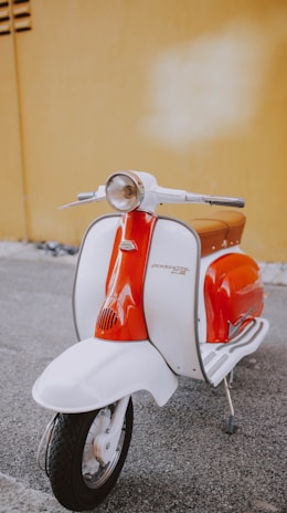 A vintage-style scooter with a vibrant orange and white color scheme is parked on a gray asphalt surface. It features a classic round headlight and chrome handlebars, set against a mustard-yellow wall.