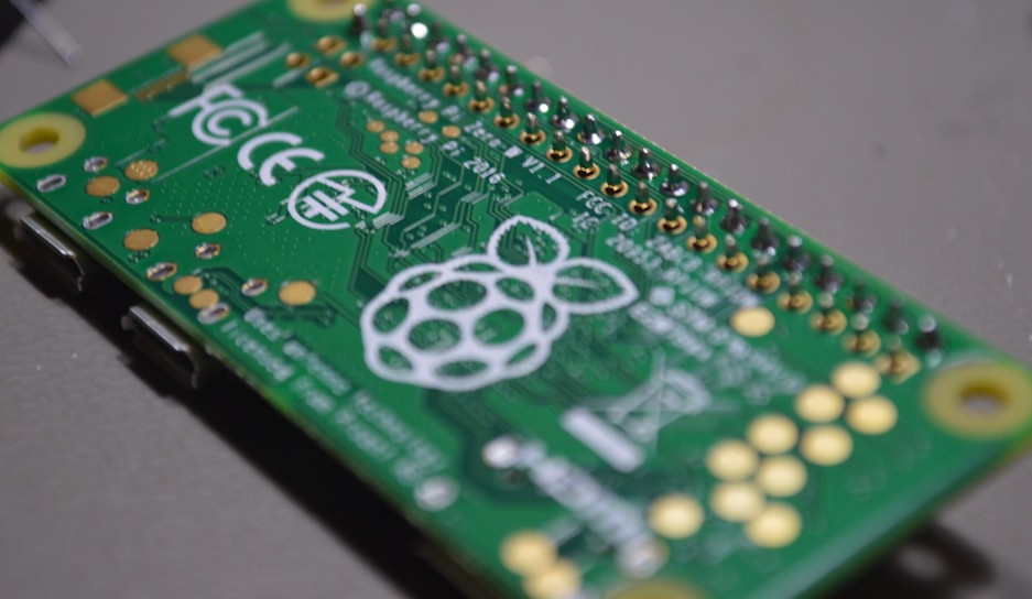 A close-up view of a green printed circuit board featuring metallic connectors and soldered points. The board has the Raspberry Pi logo prominently displayed in white along with other certifications and inscriptions.