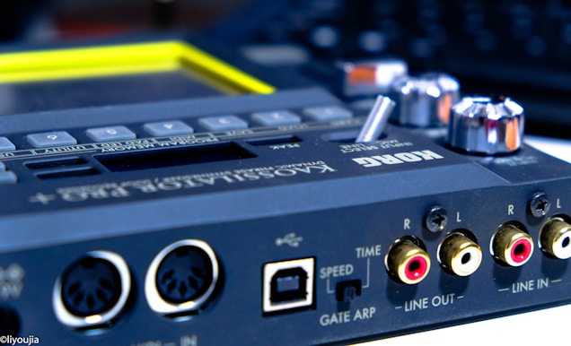 A close-up view of an electronic music device with multiple input and output ports. It features dials, buttons, and connection jacks labeled for various functions. The brand name is visible on the surface, and the lighting emphasizes the metallic and plastic components.