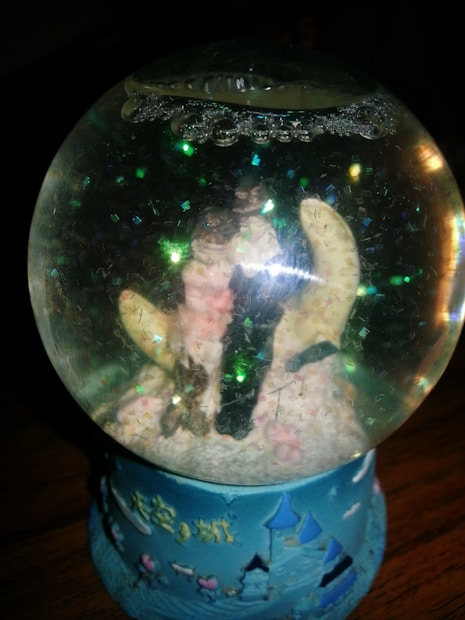 A decorative snow globe with a base painted in blue and adorned with castle and heart motifs. Inside the globe, small glitter particles are suspended, and it appears to contain a pastoral scene, though details are obscured. Bubbles are visible near the top of the globe.