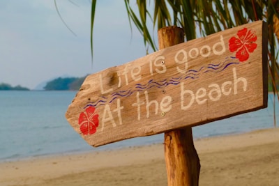 A rustic wooden sign with the words 'Life is good at the beach' painted in white, decorated with red hibiscus flowers and blue wavy lines. The sign is mounted on a wooden post and is situated on a sandy beach with the ocean visible in the background. Palm fronds hang down from the top, adding a tropical feel.