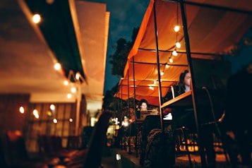 A nighttime outdoor caf&eacute; scene with string lights softly illuminating tables under a canopy. Several patrons are engaged in activities, such as using laptops, creating a cozy and focused atmosphere.