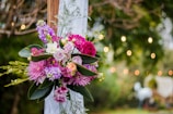 A vibrant floral arrangement featuring a variety of colorful flowers, including pink, purple, and white blooms, is set against a lush green natural backdrop. The bouquet is tied to a wooden frame with a white cloth, and soft, warm bokeh lights create a dreamy atmosphere in the background.
